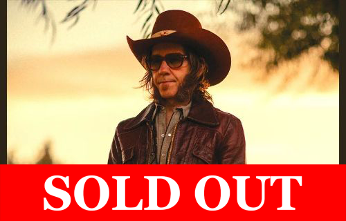 A man wearing a wide-brimmed brown hat, sunglasses, and a dark leather jacket stands outdoors in warm sunset light. He has medium-length hair and facial hair, and holds something in his hands while looking off to the side. The background is softly blurred with trees and golden sky tones.