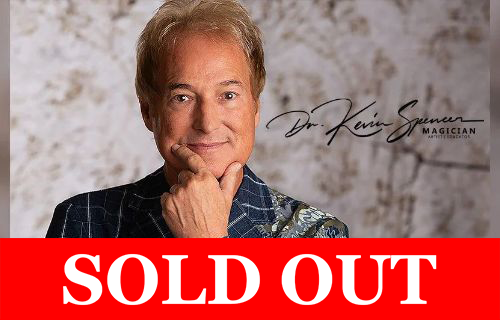 A portrait of a smiling man with light brown hair, resting his chin on his hand in a thoughtful pose. He wears a patterned blue jacket with a floral design on one shoulder. A stylized signature reading “Dr. Kevin Spencer” appears beside him, along with the word “Magician” and a sensory inclusive logo of a hand wearing headphones in the corner. A Red banner across the bottom reads "sold out" in white text.