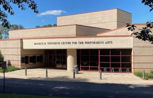 The entrance to the Maurice K. Townsend Center for the Performing Arts, featuring a curved beige facade with the building name displayed above the doorway. The entrance has glass doors and windows, a covered walkway supported by columns, and bike racks and bollards along the sidewalk. Trees frame the top of the image.
